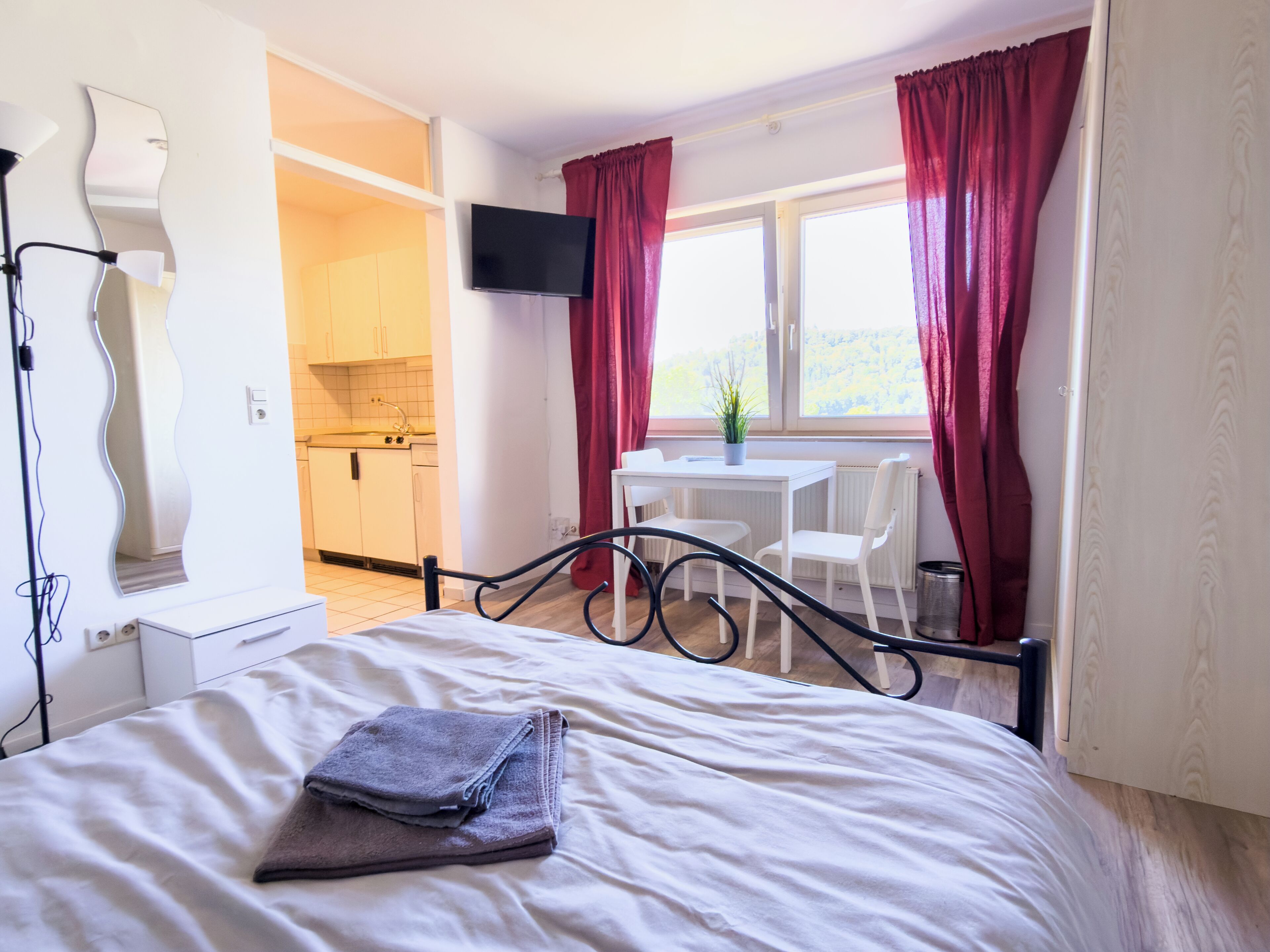 Ein Schlafzimmer mit Bett, Schreibtisch und Stuhl, ein kleiner Tisch mit Pflanze sowie Blick auf eine Küche und eine Aussicht durch ein Fenster.