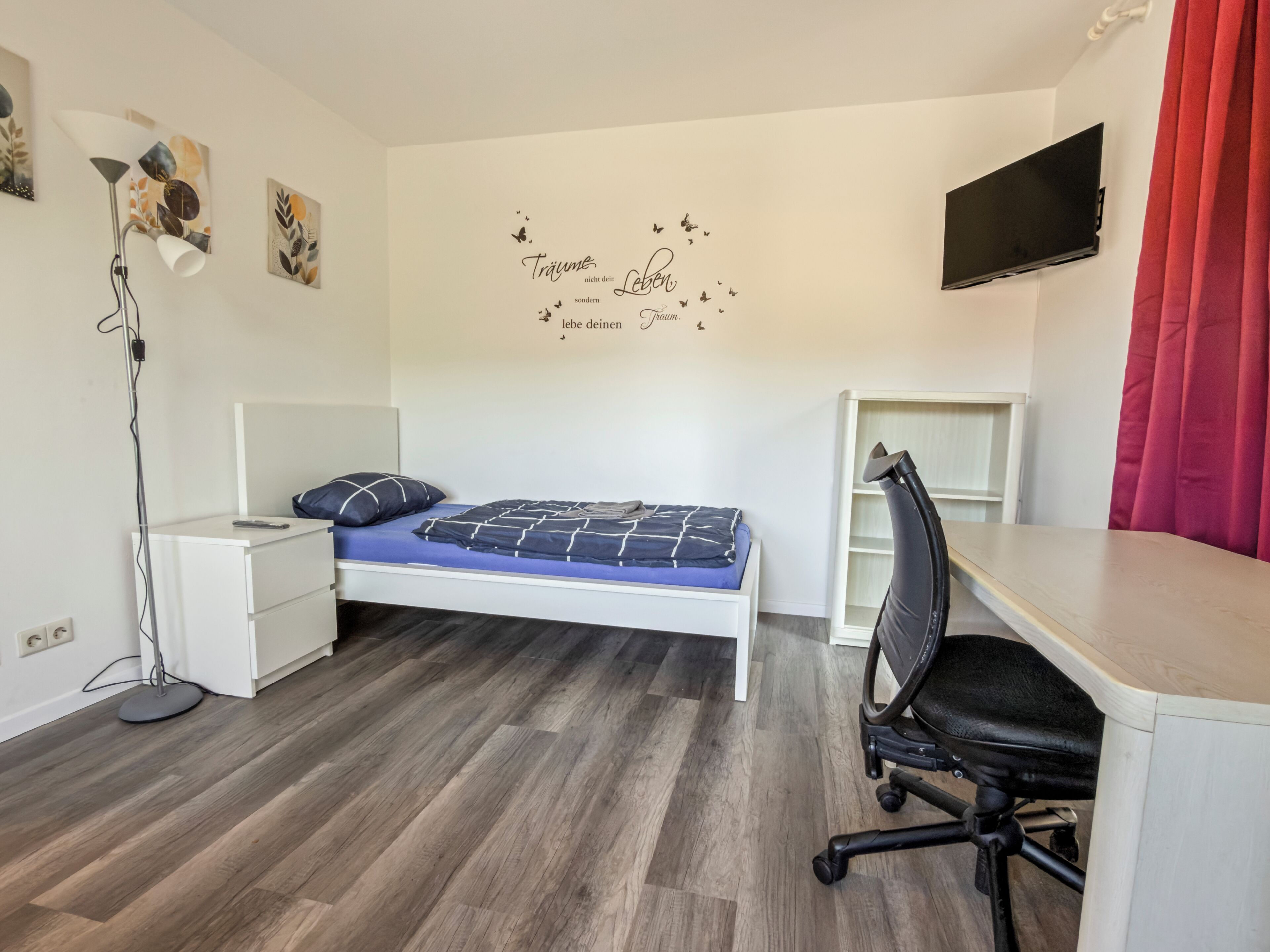 Een slaapkamer met een bed, nachtkastje, bureau, stoel en een muur met een decoratieve tekst.