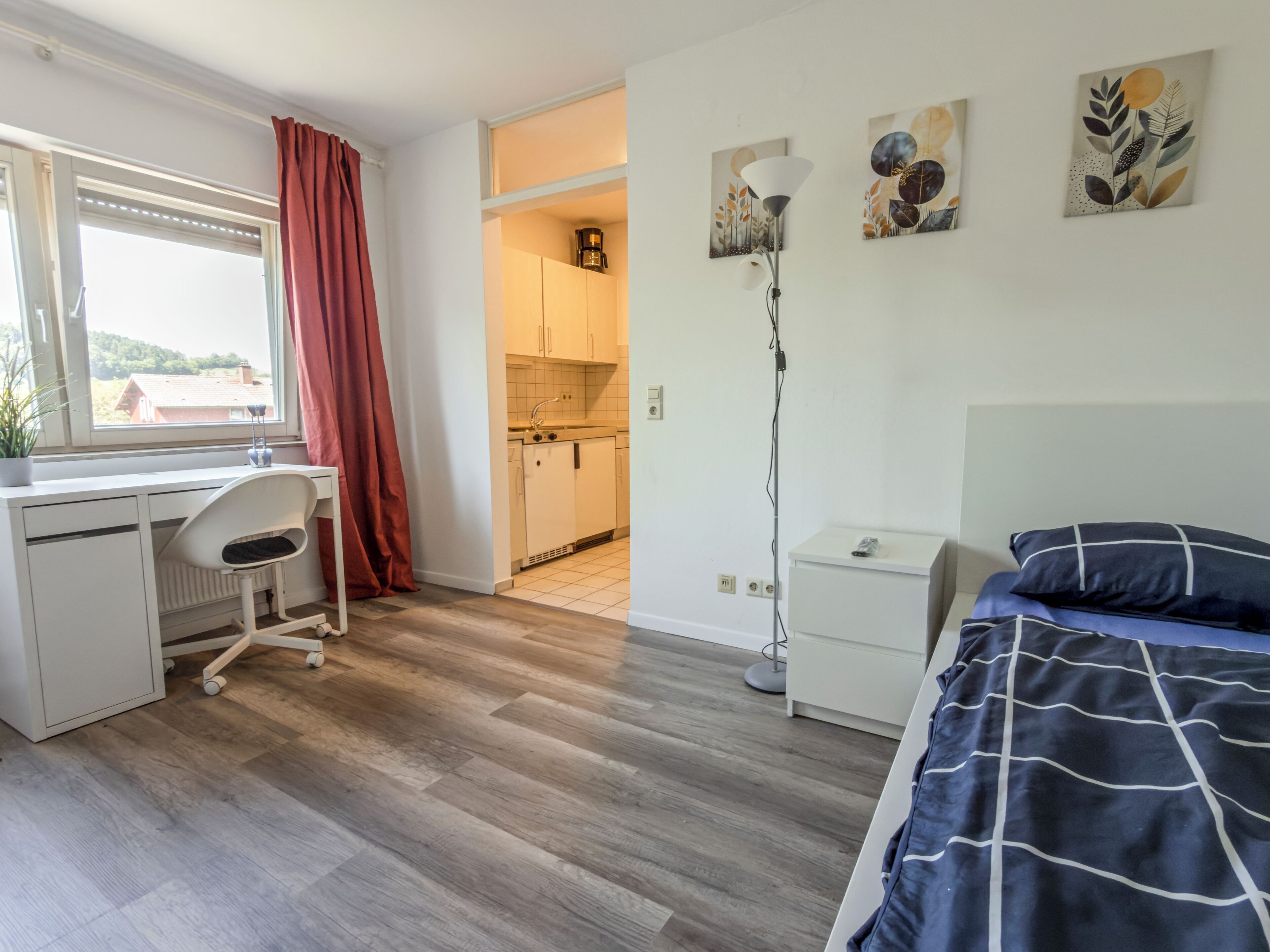 Een slaapkamer met een bed, een bureau, een stoel, een nachtkastje en een lamp. Er is een raam met gordijnen en uitzicht op een huis.