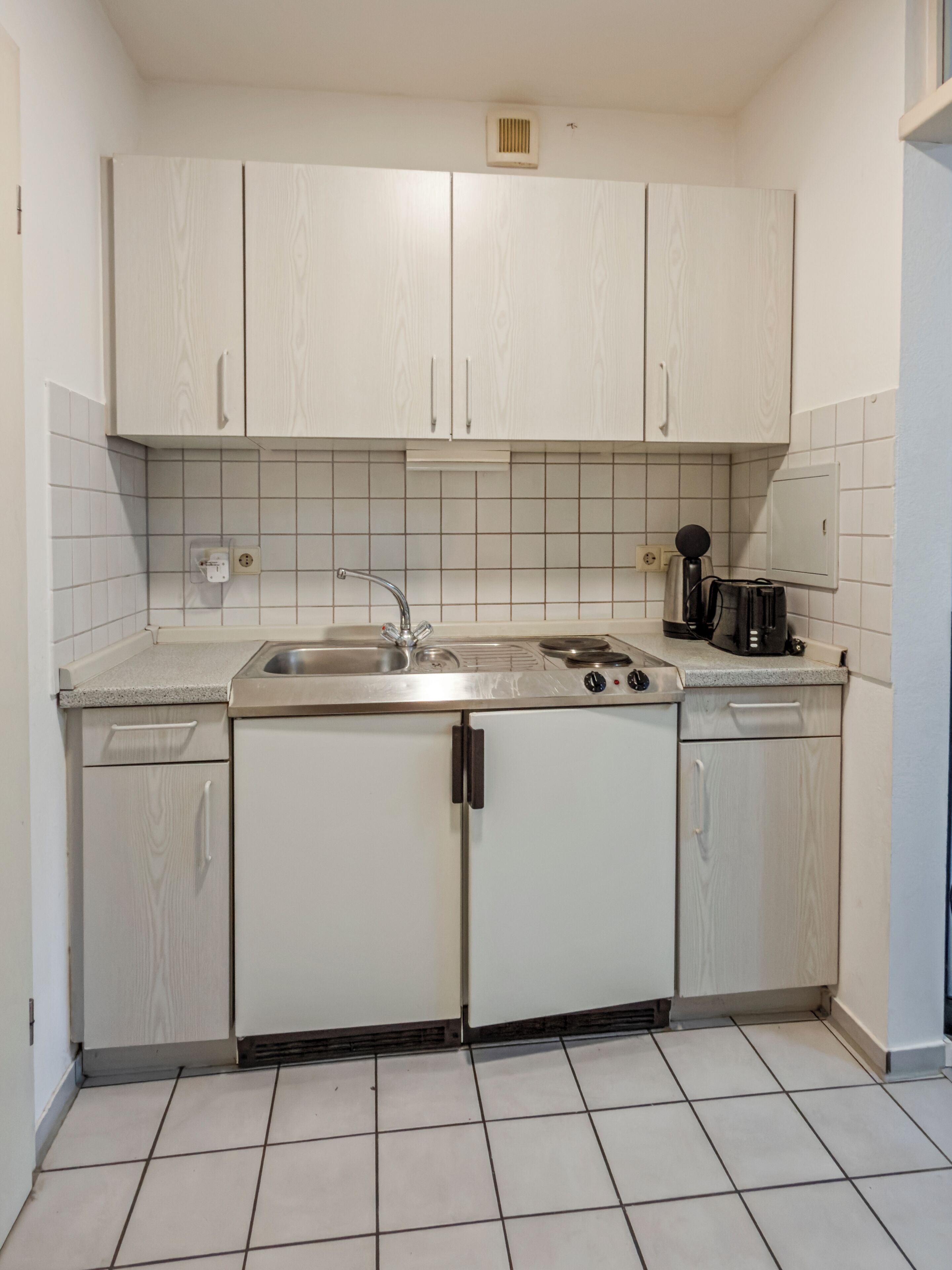 Cocina compacta con armarios blancos, un lavamanos de acero inoxidable y una cafetera.