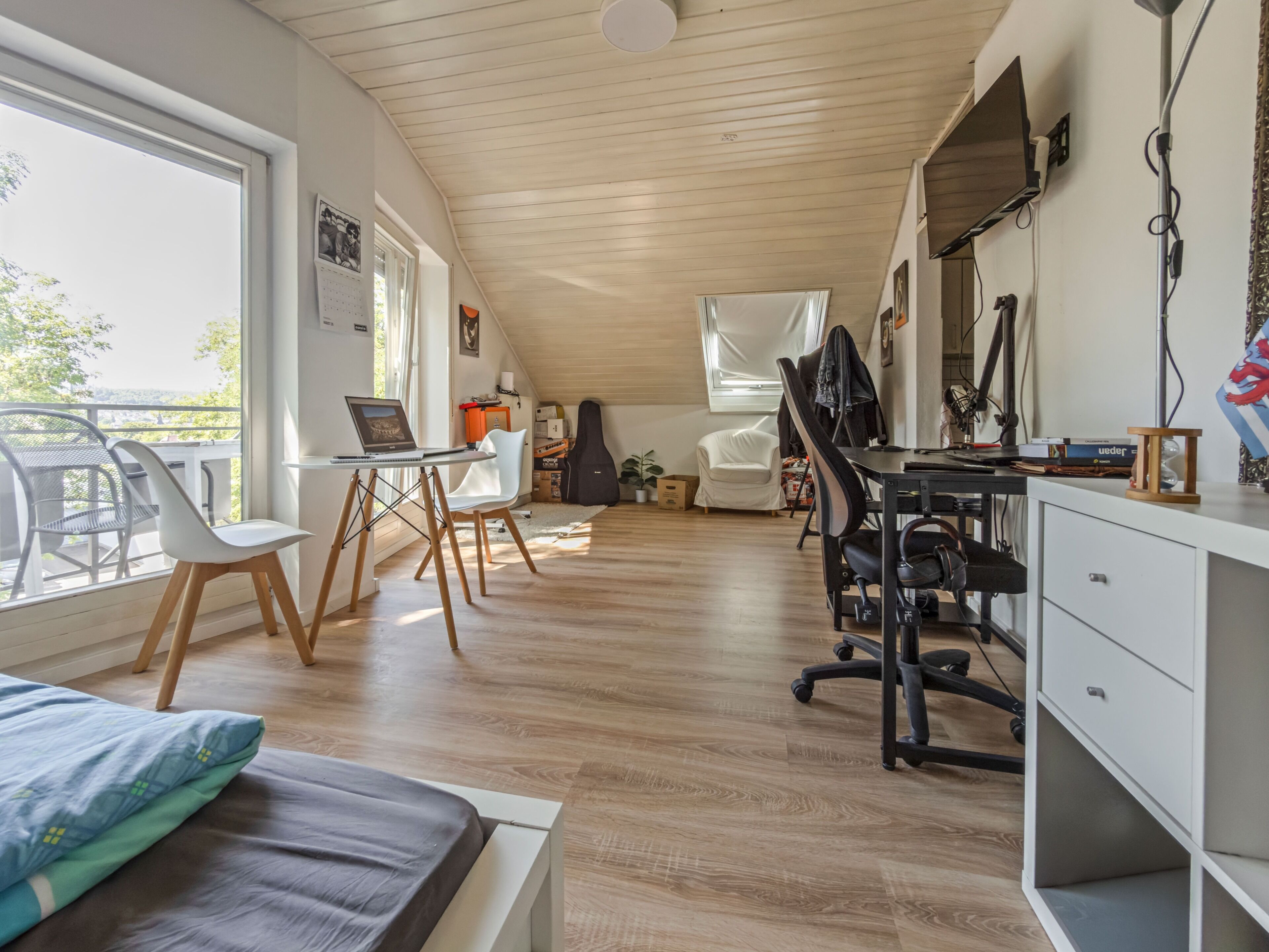 Ein modernes Zimmer mit schrägem Dach, einem Schreibtisch mit Computer, einem Stuhl, einem Bett und einem Fenster mit Blick auf einen Balkon.