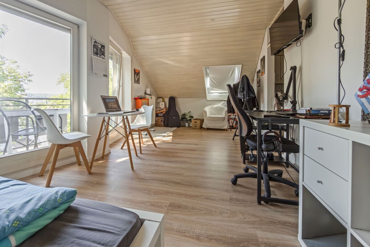 A modern room with a sloped ceiling, a desk with a computer, a chair, a bed, and a window overlooking a balcony.