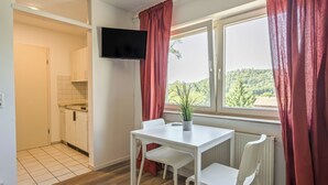 A room with a white table and chairs, a potted plant, a TV mounted on the wall, and a window with curtains.
