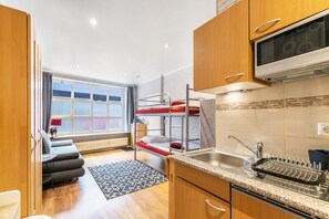 Een compacte hotelkamer met stapelbedden, een kitchenette, een zithoek en een groot raam.