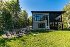 Une maison moderne à deux étages, dotée d’une grande fenêtre, d’un patio avec mobilier d’extérieur et donnant sur un paysage boisé.