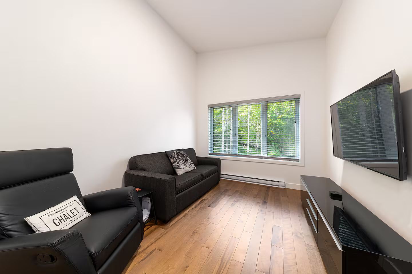 Una sala de estar moderna con un sofá y un sillón negros, un televisor de pantalla plana montado en la pared y una ventana con persianas.