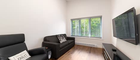 Una sala de estar moderna con un sofá y un sillón negros, un televisor de pantalla plana montado en la pared y una ventana con persianas.