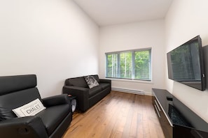 Ein modernes Wohnzimmer mit einer schwarzen Couch, einem schwarzen Sessel, einem flachbildfernseher an der Wand und einem Fenster mit Jalousien.