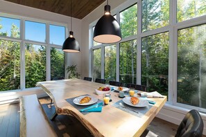 Ein Essbereich mit einem großen Tisch, zwei Pendelleuchten und Blick auf Bäume durch große Fenster.