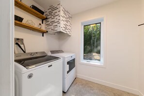 Une buanderie équipée d’un lave-linge et d’un sèche-linge à chargement frontal, avec des étagères en bois au-dessus et une fenêtre donnant sur les arbres.