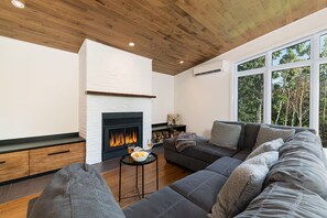 Ein modernes Wohnzimmer mit Kamin, einer großen, grauen Eckcouch und einem großen Fenster mit Blick auf Bäume.