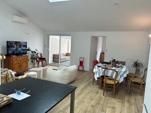 Una sala de estar moderna con un televisor de pantalla plana, un comedor con mesa y sillas, y un amplio ventanal corredera que da a un balcón.