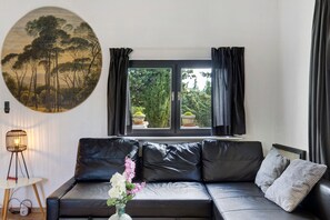 Ein modernes Wohnzimmer mit einem schwarzen Ledersofa, einem runden Wandbild, einem Fenster mit Vorhängen und einem kleinen Tisch mit einer Lampe.