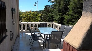 Een terras buiten met een tafel en stoelen, een witte balustrade en uitzicht op bomen en heuvels.
