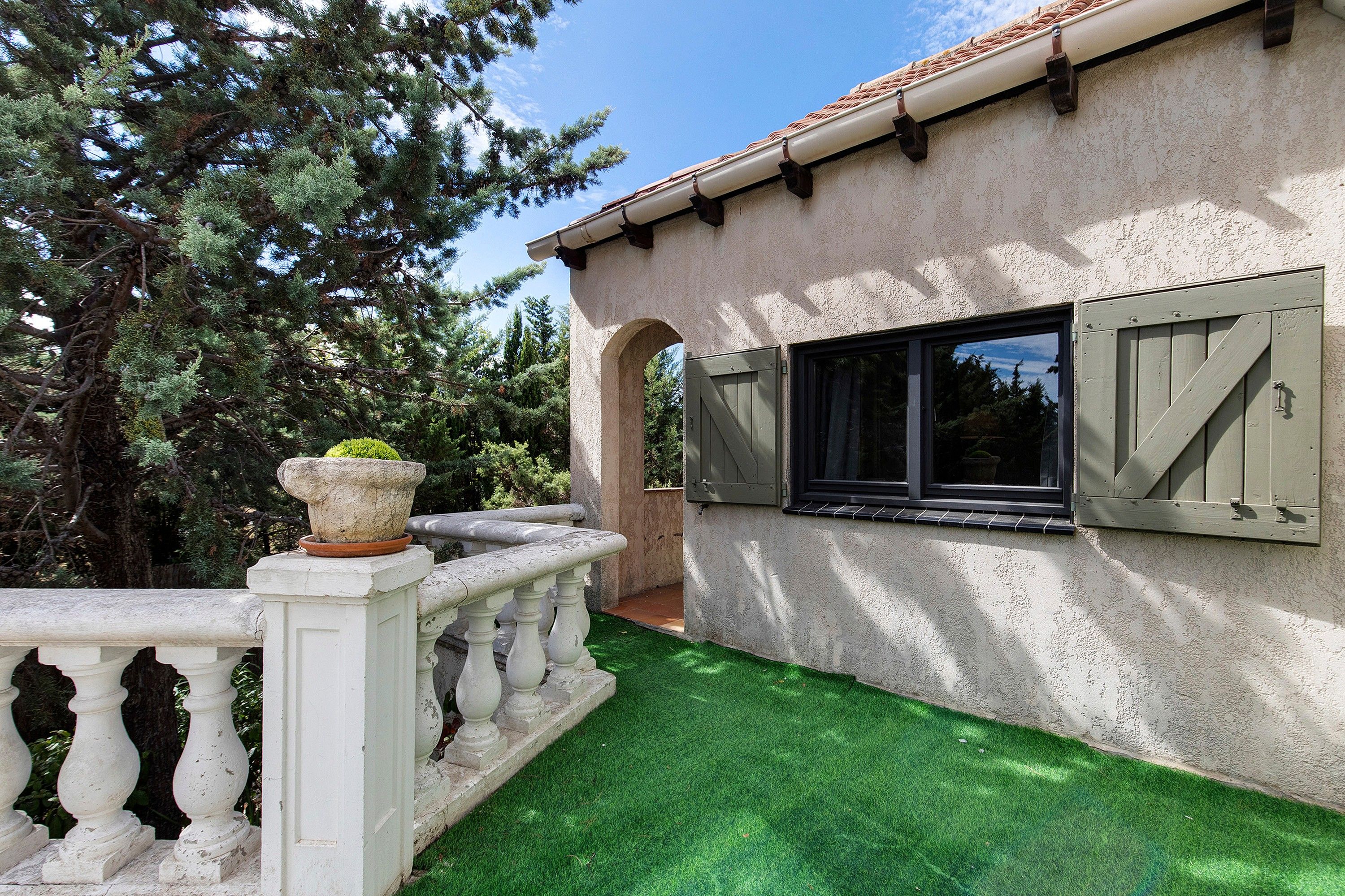 Una casa con fachada blanca, un pequeño jardín con césped artificial, una jardinera de piedra con una planta y un ventanal con contraventanas.