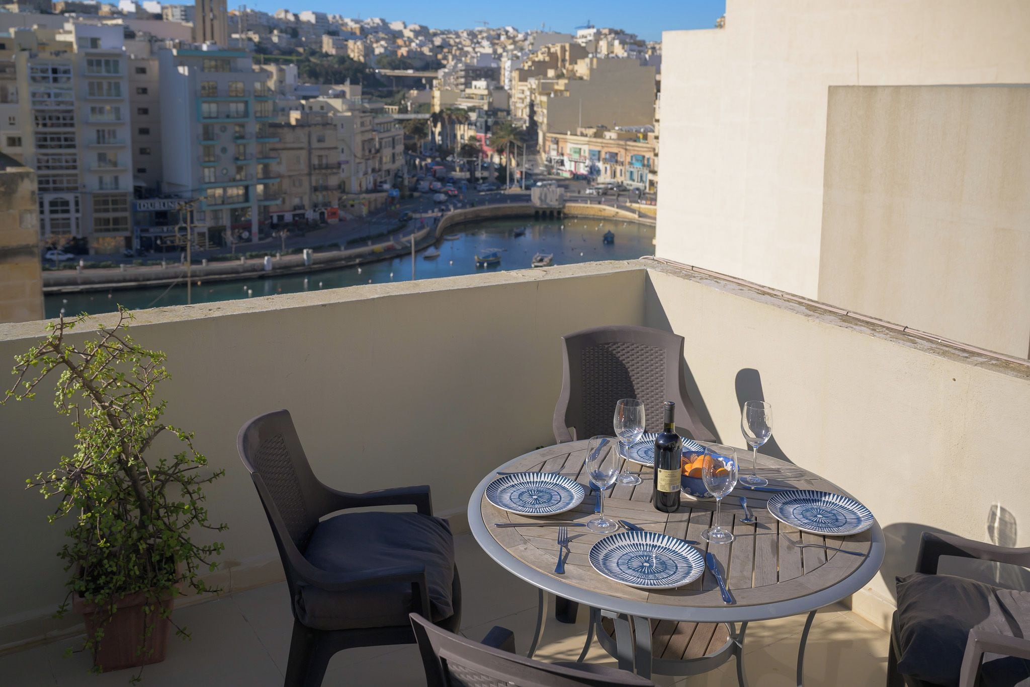 Une table ronde, entourée de chaises, et un pot de fleurs sont placés sur un balcon donnant sur une vue panoramique de la ville, avec un plan d’eau en contrebas.