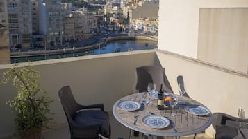Une table ronde, entourée de chaises, et un pot de fleurs sont placés sur un balcon donnant sur une vue panoramique de la ville, avec un plan d’eau en contrebas.