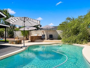 A swimming pool with lounge chairs and umbrellas, surrounded by a stone wall and greenery. - Peoria Home | Pool | Game Room (Peoria)