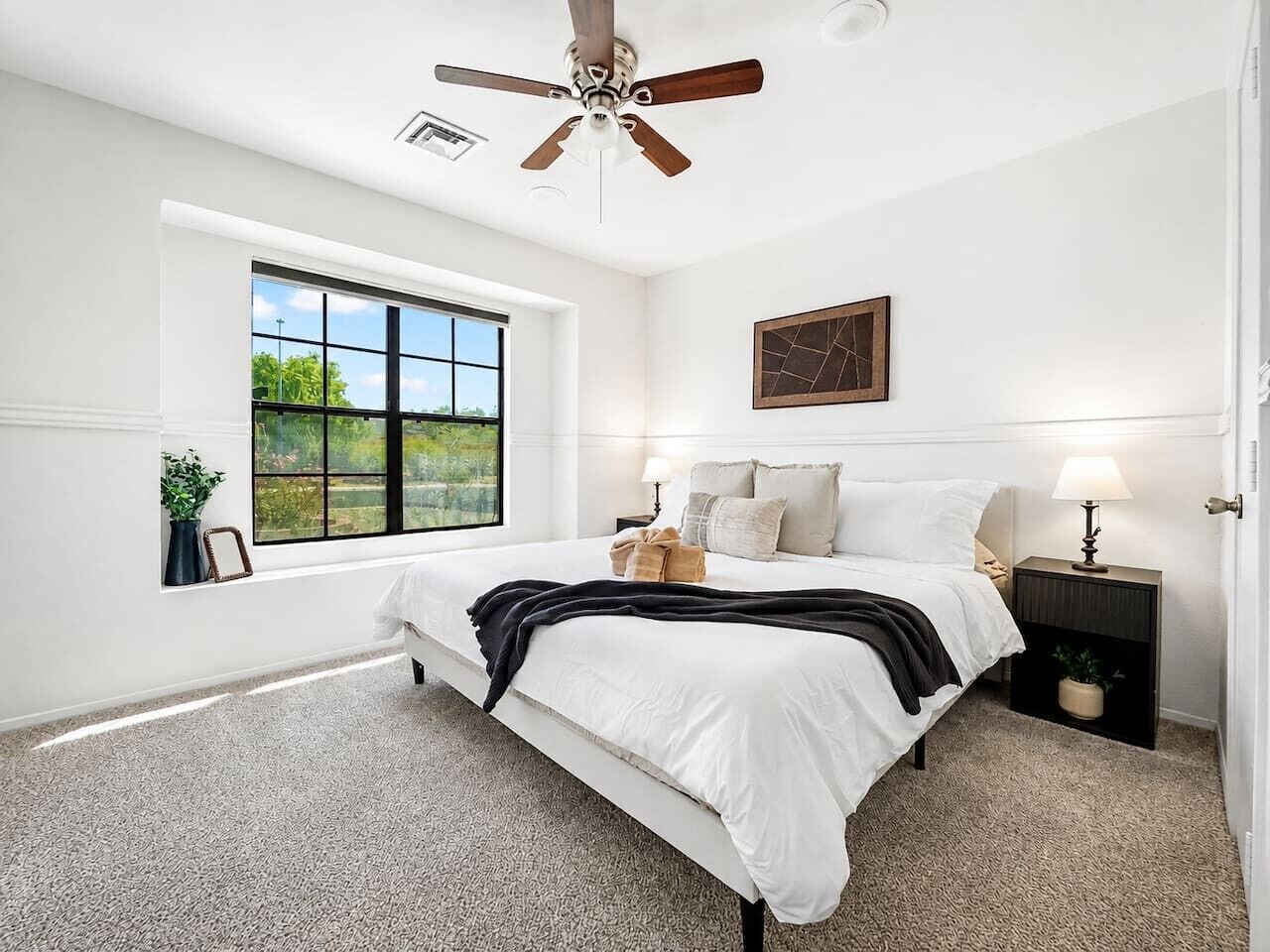 A bedroom with a large window, a ceiling fan, a bed with white bedding, a bedside table with a lamp, and a wall-mounted artwork.