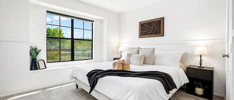 A bedroom with a large window, a ceiling fan, a bed with white bedding, a bedside table with a lamp, and a wall-mounted artwork.