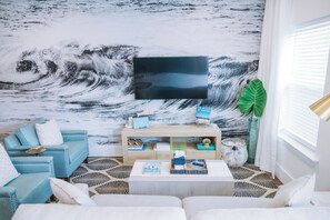 A living room with a large wave mural, a TV, a blue sofa, a coffee table, and a plant. - The Cove Reunion HQ - Sleeps 22 - Gym - Game Room - Theater - Spa (Santa Rosa Beach)
