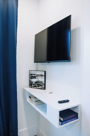 A room with a mounted flat-screen TV, a framed picture of a vintage car, a remote control, and a shelf with books. - The Cove Reunion HQ - Sleeps 22 - Gym - Game Room - Theater - Spa (Santa Rosa Beach)
