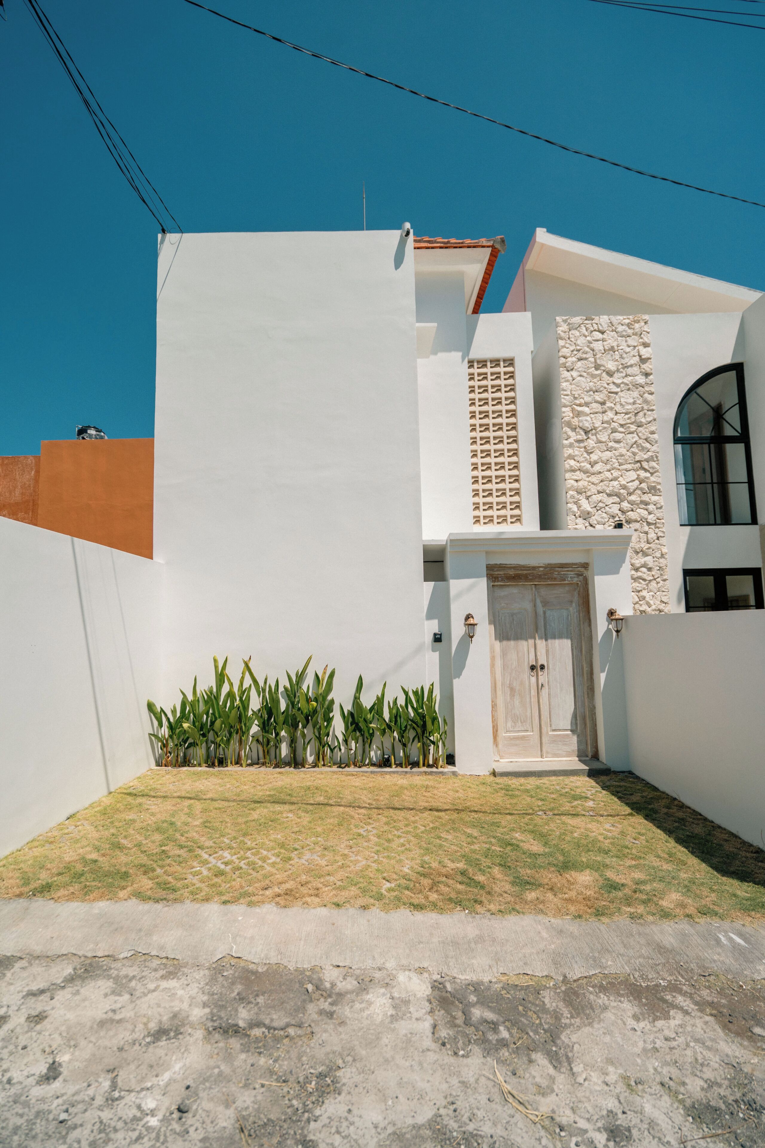 Una casa moderna de dos pisos con fachada blanca, un muro de piedra decorativo, una puerta de madera y un pequeño jardín con plantas.