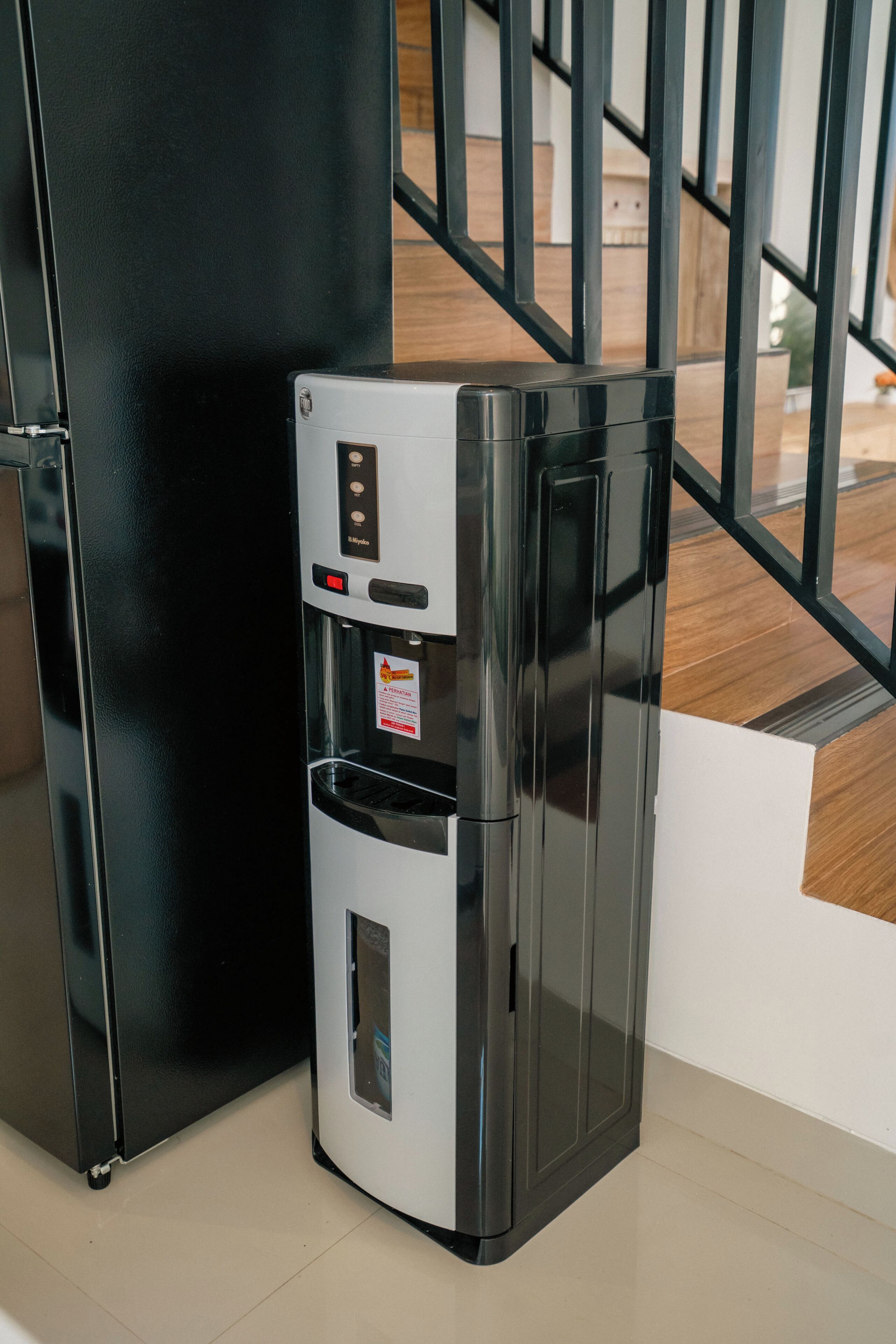 Un refrigerador compacto y moderno con panel de control digital, ubicado junto a un pilar negro en una habitación con piso de madera y una escalera al fondo.