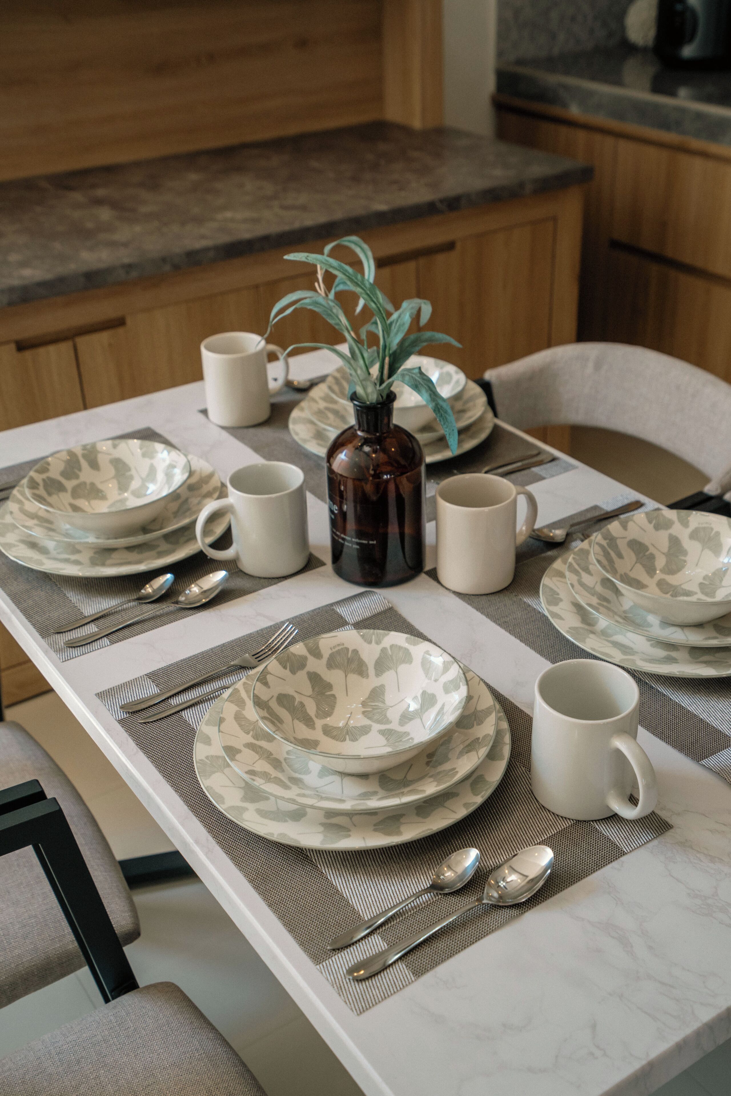 Una mesa de comedor dispuesta con platos, tazas y cubiertos, con un florero de plantas verdes en el centro.