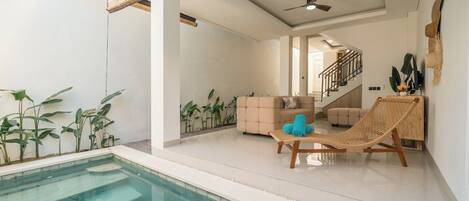 A modern indoor pool area with a lounge chair and a small table, surrounded by plants and a staircase leading to an upper level.