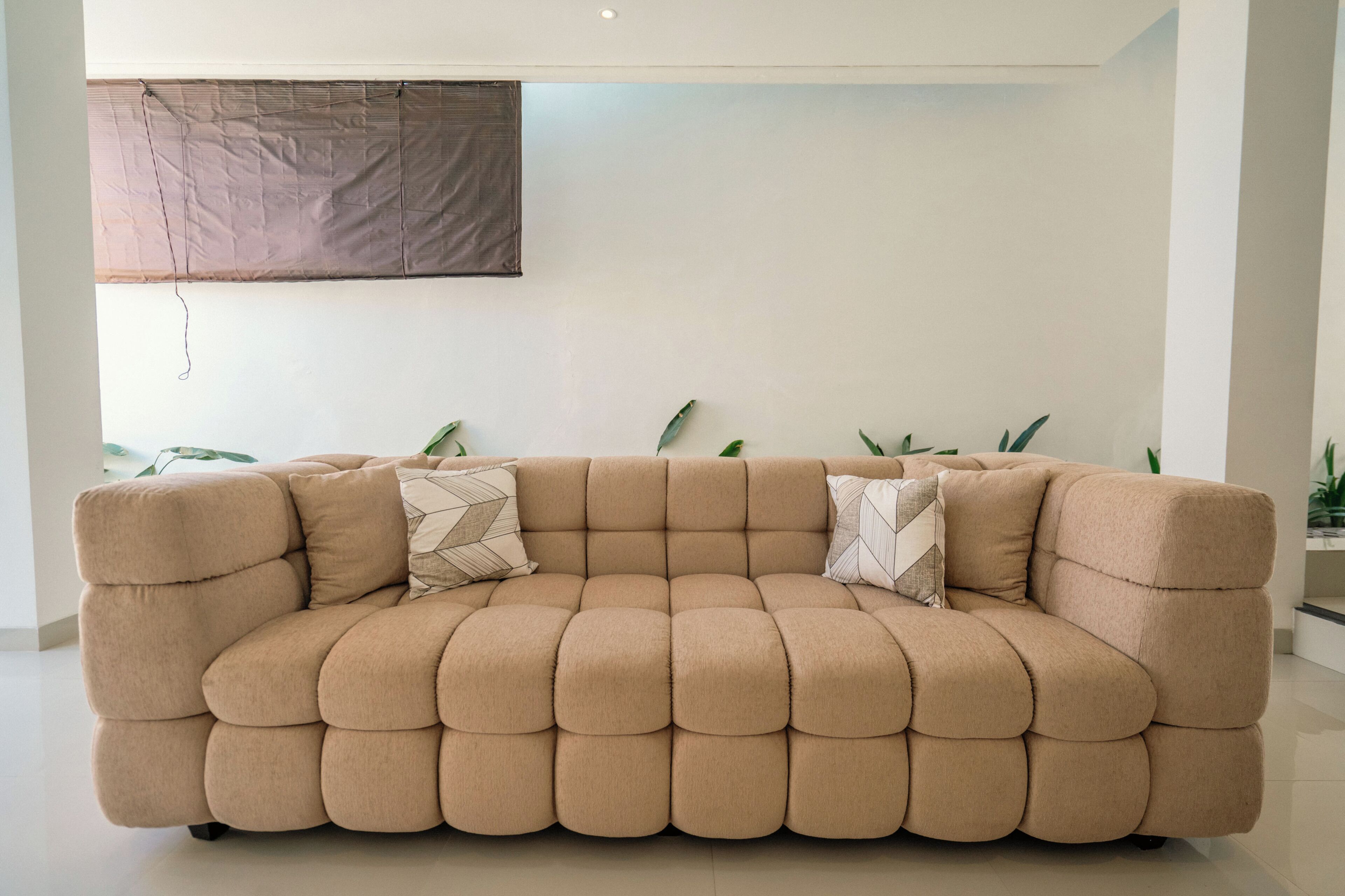 Un sofá seccional beige con un cojín de estampado geométrico, ubicado en una habitación con pared blanca lisa y una ventana con persiana marrón.