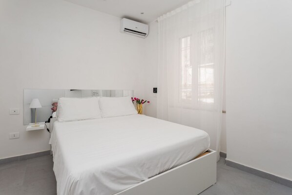 Een slaapkamer in het wit met een bed, nachtkastje, lamp, een vaas met bloemen en een raam met gordijnen.
