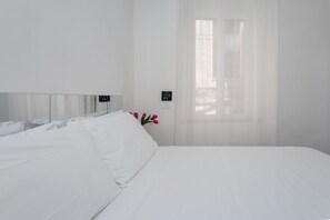 Een hotelkamer met een bed, een nachtkastje, een raam met dunne gordijnen en een bedieningspaneel aan de muur.
