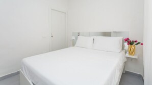 Minimalistinen hotellihuone, jossa on sänky, yöpöytä, lamppu ja maljakko, jossa on kukkia.