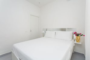 Een minimalistisch hotelkamer met een bed, een nachtkastje, een lamp en een vaas met bloemen.