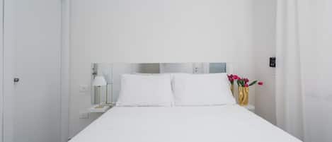 Une pièce blanche avec un lit, une table de chevet, une lampe et un vase rempli de fleurs.