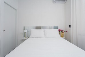 Een witte kamer met een bed, een nachtkastje, een lamp en een vaas met bloemen.