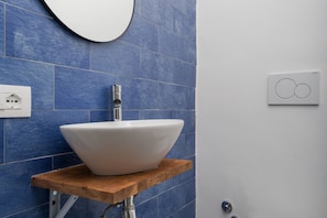 Een badkamer met een witte wasbak op een houten plank, blauwe tegels aan de muur, een ronde spiegel en een lichtschakelaar.