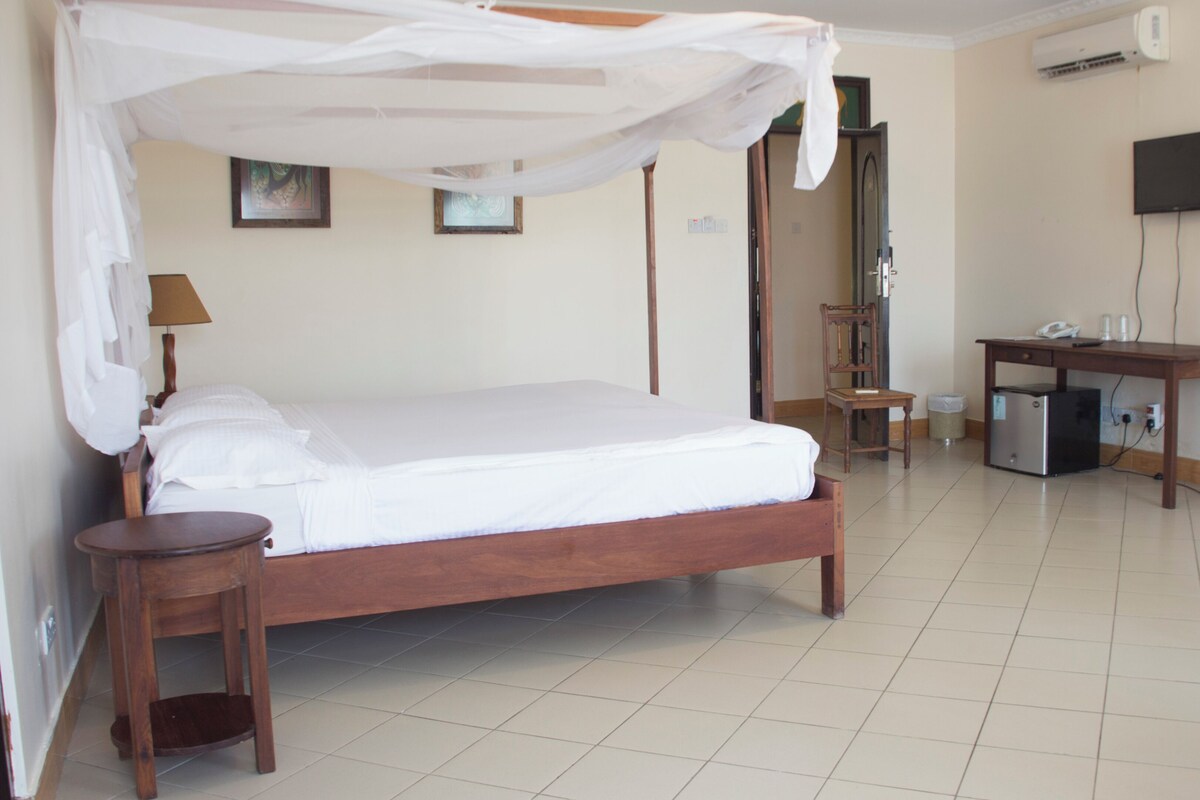 A hotel room with a four-poster bed, a desk with a chair, a television, and an air conditioning unit.
