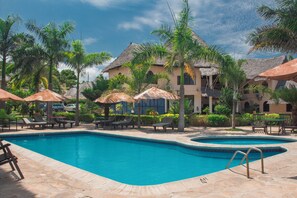 A swimming pool area with lounge chairs and umbrellas, surrounded by palm trees and a building with a thatched roof. - Millennium Sea Breeze Resort (Pwani Bagamoyo)