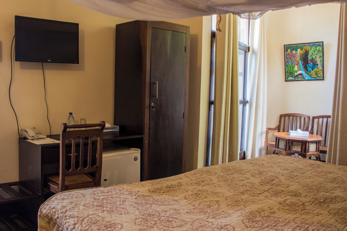 A bedroom with a bed, a desk, a chair, a wardrobe, a television, and a window with curtains.