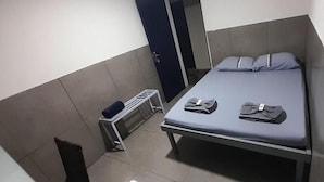 Un llit individual amb matalàs i coixins grisos, una petita taula de nit i un radiador fixat a la paret.