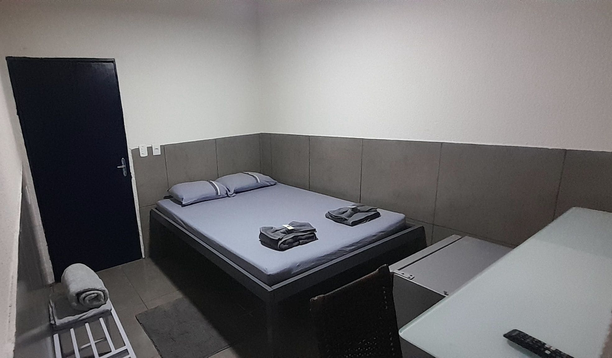 A small, single-bed room with a grey headboard, a bedside table, and a remote control on the floor.
