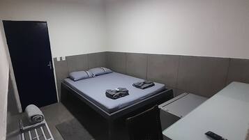 A small, single-bed room with a grey headboard, a bedside table, and a remote control on the floor.