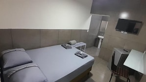 Habitació amb un llit individual, matalàs gris, almoines, taula petita, cadira, televisió penjada a la paret i un lavabo a la cantonada.