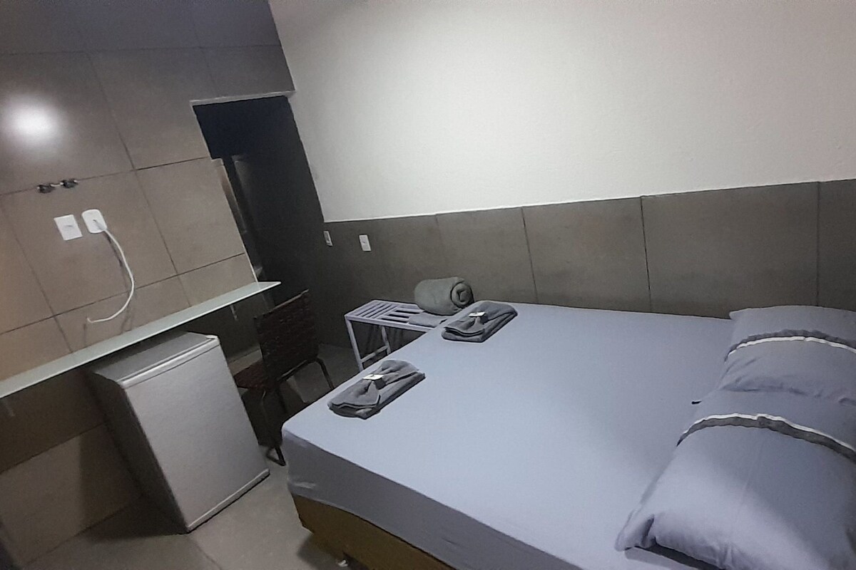 A small, compact hotel room with a bed, a desk, a chair, a small refrigerator, and a shower area.