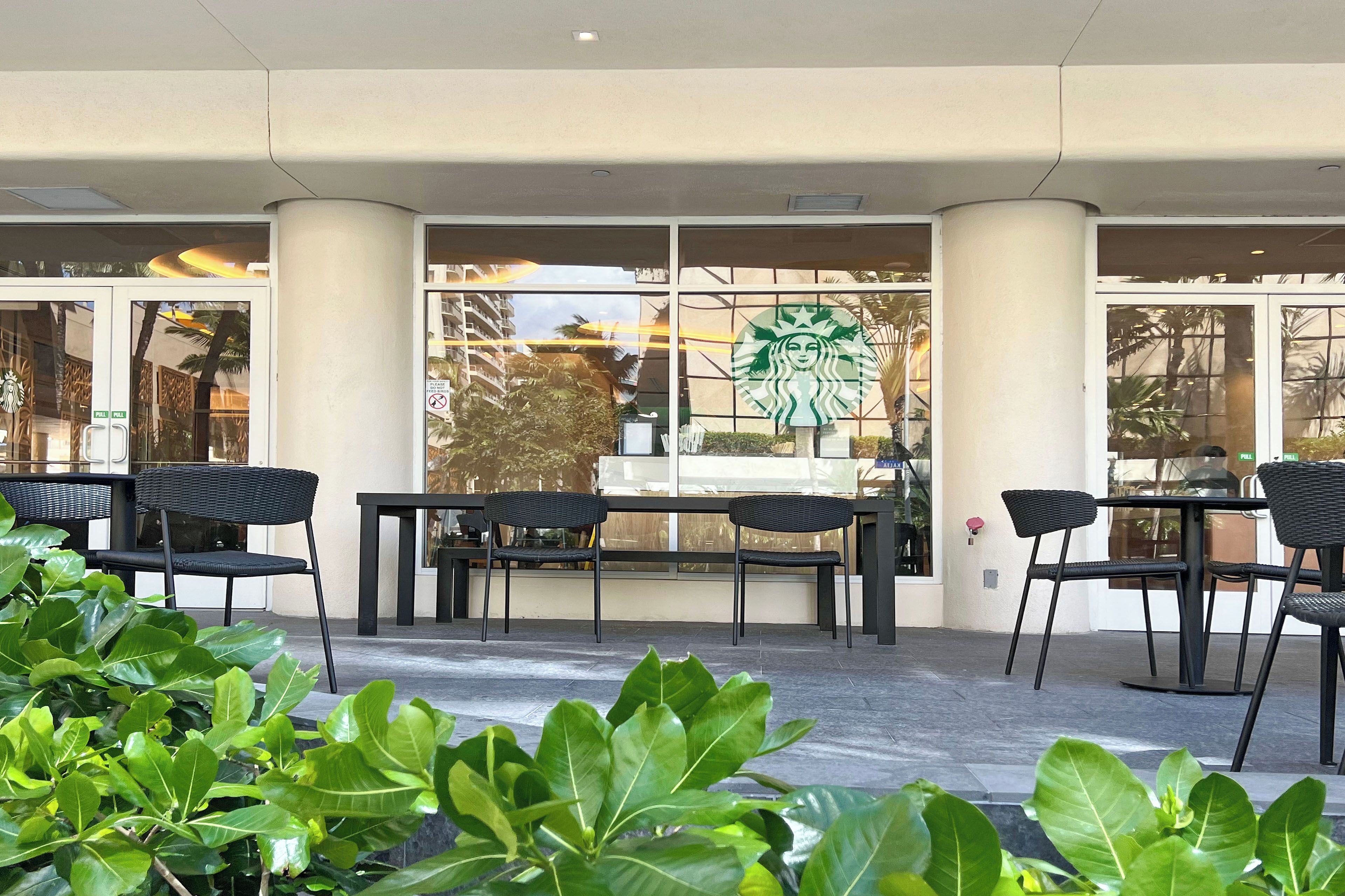 Ein Außenbereich mit schwarzen Stühlen und Tischen, einem Starbucks-Logo am Fenster und Pflanzen im Vordergrund.