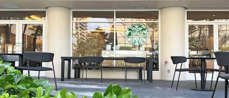 Ulkoterassi, jossa on mustia tuoleja ja pöytiä, Starbucks-logo ikkunassa ja vihreitä kasveja etualalla.
