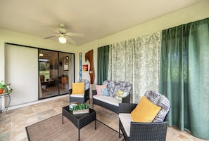 A sunlit room with a ceiling fan, a sofa set with patterned cushions, a small table with a plant, and a view through a glass door into another room.