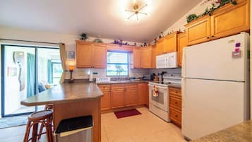 Une cuisine avec des meubles de rangement en bois, un réfrigérateur blanc, un four à micro-ondes et une cuisinière. Il y a une fenêtre avec des stores et une porte qui donne sur une autre pièce.